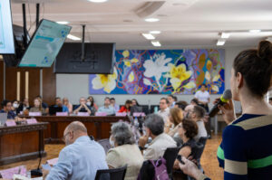 A imagem mostra a sala de reunião do CONSUNI, com diversos participantes, e em primeiro plano, a Coordenadora de Planejamento e Gestão Estratégica apresentando o PLS